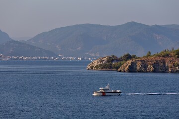 Obraz premium A boat is sailing on blue water near a rocky coast. Mountains are visible in the distance with a hazy sky overhead in Icmeler - Marmaris - Mugla - Turkey