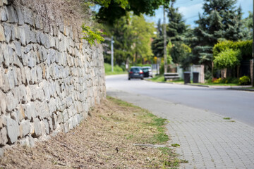 Zabytkowy, kamienny mur oporowy wykonany z nieregularnych, zwietrzałych kamieni, zabezpieczający zbocze malowniczej miejskiej ulicy. Szorstka faktura muru kontrastuje z gładkim, jasnym chodnikiem. 