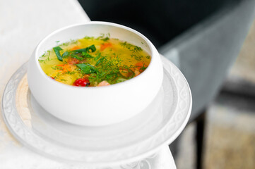 White bowl of vegetable soup garnished with fresh herbs, served on a white plate over patterned tablecloth, with soft-focus background