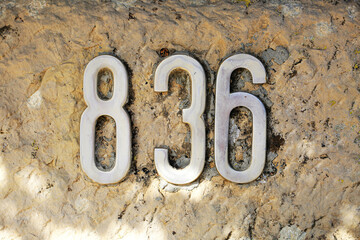 House number 836 displayed with metallic numerals mounted on a natural stone wall. Silver digits contrast with rustic textured rock, concept of address signage, property marker, and architecture