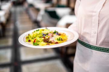 Waiter holding a plate with colorful gourmet salad in restaurant setting, featuring leafy greens, bell peppers, and edible flowers.