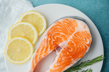 Closeup raw salmon steak with lemon slices and rosemary on white plate over blue background