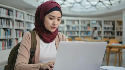 Muslim female student in hijab using laptop in modern library, young woman studying and smiling while researching online, academic diversity and education technology concept