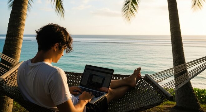 A man in a white t-shirt lounges in a hammock by the ocean, using a laptop.
