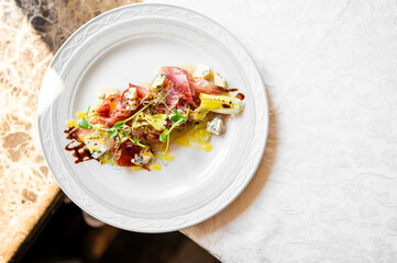 Gourmet appetizer with prosciutto, blue cheese, greens, balsamic glaze, and olive oil on elegant white plate over marble and linen table setting