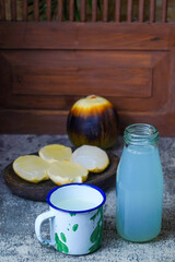 A refreshing bottle of legen or palmyra palm sap and a classic enamel mug, with palmyra fruit and kernels blurred in the background on a textured gray surface.