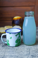 A refreshing bottle of legen or palmyra palm sap and a classic enamel mug, with palmyra fruit and kernels blurred in the background on a textured gray surface.