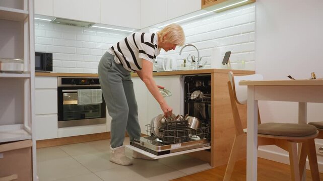 An elderly woman unloads the dishwasher with clean kitchenware in her home kitchen. She puts away plates and pots into the cabinet, performing a daily household chore for a tidy house.