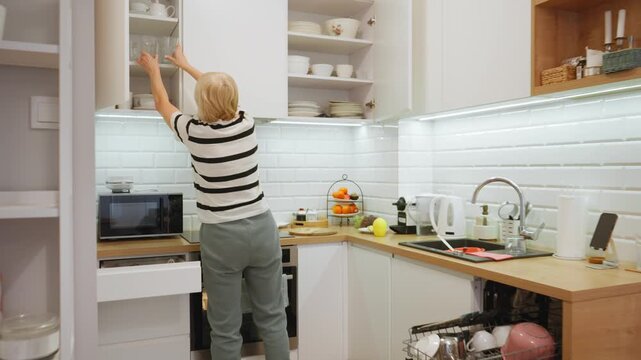 A housewife puts clean dishes away after using the dishwasher. She organizes the kitchen space by neatly arranging mugs and glasses on the shelf, keeping her home tidy and clean.