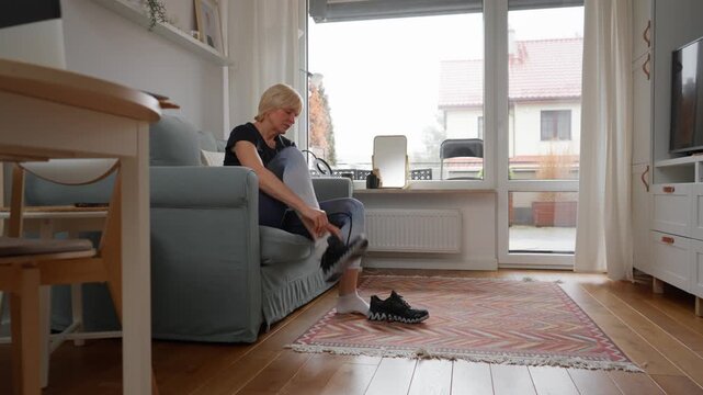 An active senior woman gets ready for her fitness training in the living room. She puts on sportswear and ties the laces on her sneakers, motivated for a run, walk, or workout.