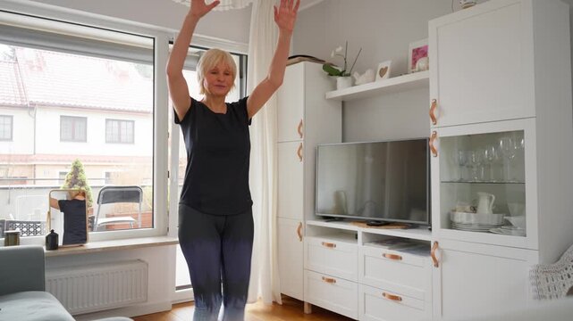 A senior woman follows an online fitness tutorial on her laptop for a home workout. She is taking a virtual lesson with a remote coach, using technology for e-learning and staying active.