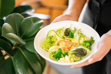 Fresh shrimp salad with cucumber, broccoli, leafy greens, and diced fruit in a white bowl, held by hands with a green plant in the background