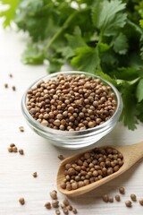 Coriander seeds in bowl, spoon and fresh cilantro leaves on light wooden table, closeup