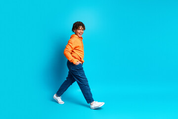 Cheerful young boy wearing a bright orange hoodie and blue jeans walks confidently against a vibrant blue background.