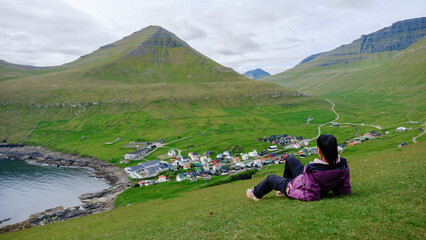 Naklejka premium Nestled in the lush green hills of the Faroe Islands, an Asian woman person relaxes, gazing at the picturesque Gjogv village and serene coastline.