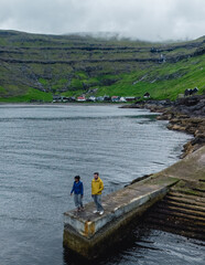 Two adventurers explore the picturesque harbor of the Faroe Islands, surrounded by stunning cliffs and lush green hills. Tjornuvik village