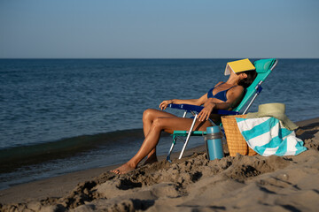Woman in blue bikini napping on beach chair with yellow book covering her face, relaxing by the calm sea with straw hat, basket, towel, and thermos on sunny summer day