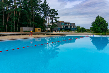 Modern swimming pool at Słupca Lake in Poland, with beach, sun loungers and buildings in the background, surrounded by trees and greenery under a cloudy sky.