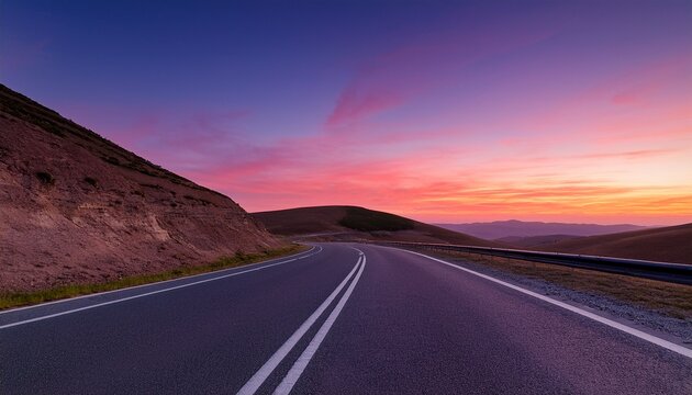 an empty road at twilight with the sky filled with warm hues of pink and purple the paved highway stretches into the distance curving around hills under a clear sky