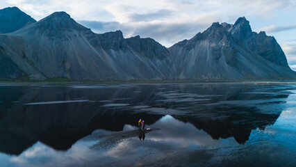 Stokksnes showcases the stunning Vestrahorn mountain, where two adventurers explore the black sand beaches as the reflection creates a mirror-like effect on the calm waters at dusk in Iceland.