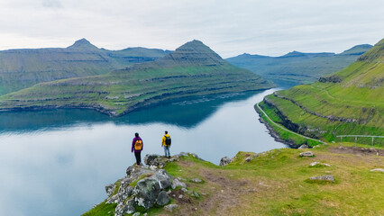 Explore the majestic landscapes along the Gonguturur Hvithamar Trail in the Faroe Islands. Two hikers stand at the cliffs edge, surrounded by lush green hills and tranquil waters reflecting the sky.