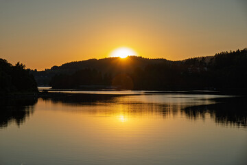 Sunset behind forested hills with glowing sun rays reflecting on still lake surface, warm golden evening light on horizon.