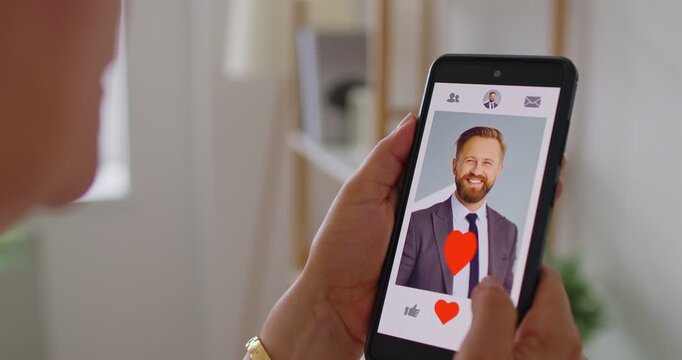 Woman browsing dating app on smartphone and pressing heart icon on man&rsquo;s smiling profile. Online dating interaction. She shows romantic interest. Bright indoor background scene.