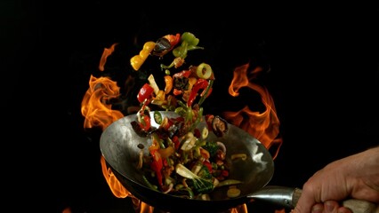 Closeup of chef throwing asian vegetable with meat mix from wok pan in fire. Fresh asian food preparation on black background.