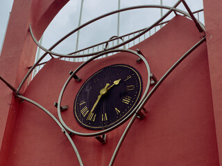 A detailed view of a large, black and gold clock with Roman numerals, set into a striking red wall and framed by modern metal structures.