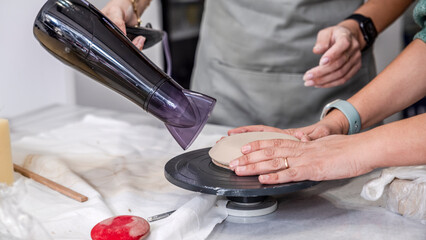 In an art studio with an environment adorned with artistic paintings in the background, a beautiful middle-aged woman artist is drying a clay-shaped plate with a hairdryer at a table