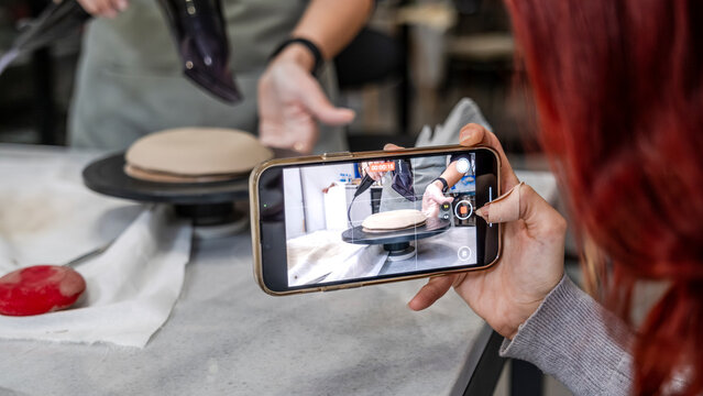 In an art studio a beautiful middle-aged woman artist is shaping clay at a table, while a young woman next to her is taking a photo with her phone - Powered by Adobe