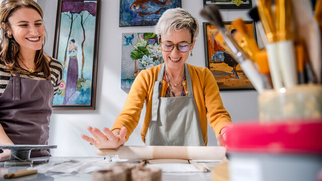 In an art studio with an environment adorned with artistic paintings in the background, two beautiful middle-aged woman artist is shaping clay with a rolling pin at a table