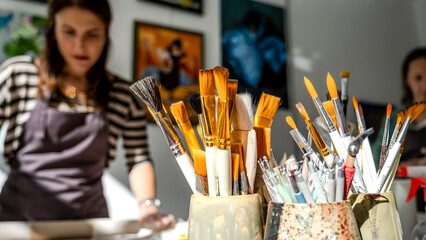 A close-up shot of paintbrushes in the foreground, with two women artists working on handmade...