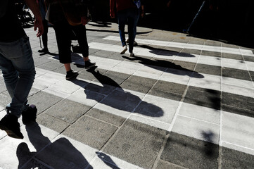 Pedestrians walking across a zebra crossing with strong sunlight casting long shadows. Urban movement and street life captured in high contrast. Concept of anonymity, transition and city rhythm.