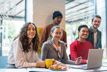Group of diverse young professionals smiling while working together in a modern cafe with laptop and coffee. Concept of teamwork, coworking, remote work and multicultural collaboration.