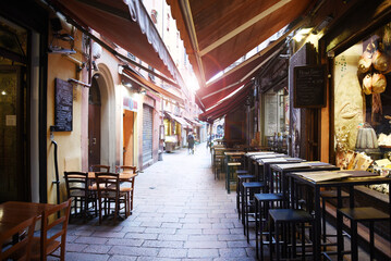Traditional Italian alley with outdoor cafés and restaurants in Bologna city center. Early morning light creates a charming atmosphere in this empty cobblestone street. Culinary tourism concept.