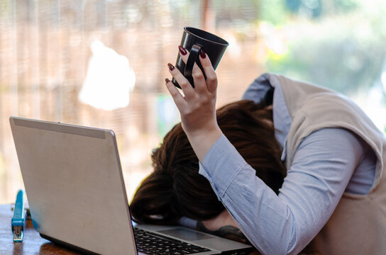Exhausted young female employee feeling fatigue lying on table and lifting cup of coffee lack of energy in morning office tired of stress problems sleeping at workplace overwork concept