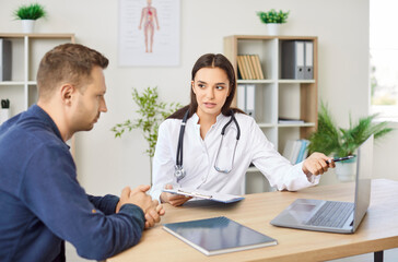 Cheerful friendly female doctor at medical office in hospital showing examination results on laptop monitor screen and giving consultation to a young man patient sitting in clinic.