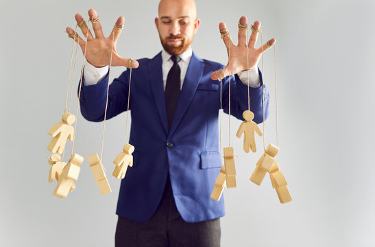 Bottom view of a young businessman puppeteer wearing suit controlling wooden human figures marionettes by hands hanging on rope isolated on white background. Manipulating and people control concept.