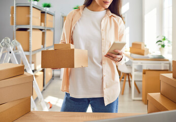 Web store owner or inventory manager working in warehouse, preparing merchandise in stock for sending, packing stuff in boxes in storage room, checking data in e commerce logistic app on mobile phone