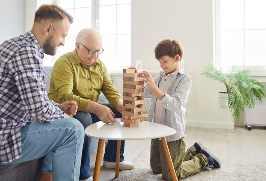 Happy multi generational family playing block removal game at home. Smiling child boy together with father and elderly grandfather playing board game in living room