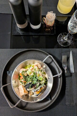 Top-down of seafood stew with mussels and salmon in creamy sauce in metal pot on black plate. Fine dining setting includes dark table, drinks and elegant cutlery, showcasing rich meal