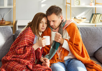 Young couple man and woman sitting on sofa at home wrapped in a blanket trying to warm up with hot cup of drink suffering from heating problems or feeling unhealthy with influenza.