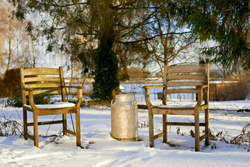 Two empty wooden chairs and a vintage milk can sit in a tranquil, snowy garden during a sunny...