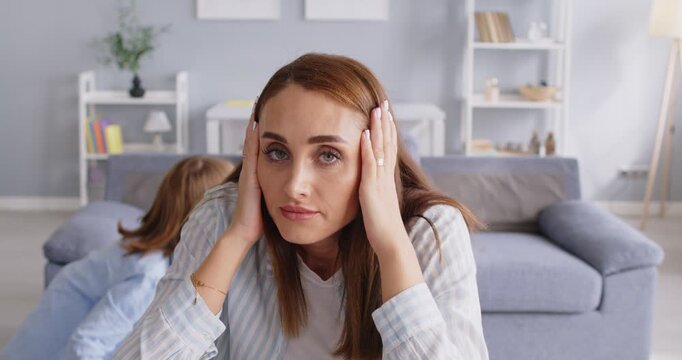 Stressed young mother suffering from her active noisy children running around and shouting. Tired parent with pained expression clutching her head, having problem with naughty kids playing loudly.