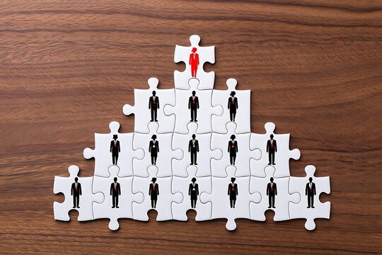 Hierarchical organizational structure represented by a pyramid of puzzle pieces with business people figures and a red leader at the top on a wooden background
