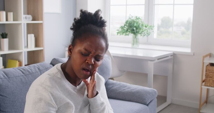Unhappy african american woman sitting at home touching her cheek in suffer from bad toothache, sick girl having terrible sudden tooth pain, needs dental treatment, stomatology, dentist visit