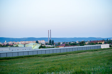 Obraz premium A grassy field stretches toward an industrial complex and townscape nestled beneath rolling hills and a clear sky.