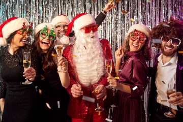 Happy young people group of friends holding champagne glasses having fun standing together with a man in Santa Claus costume celebrating New Year's Eve party. Christmas holiday concept.