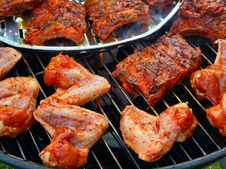 Marinated chicken wings and pork ribs are cooking on a hot outdoor barbecue grill.